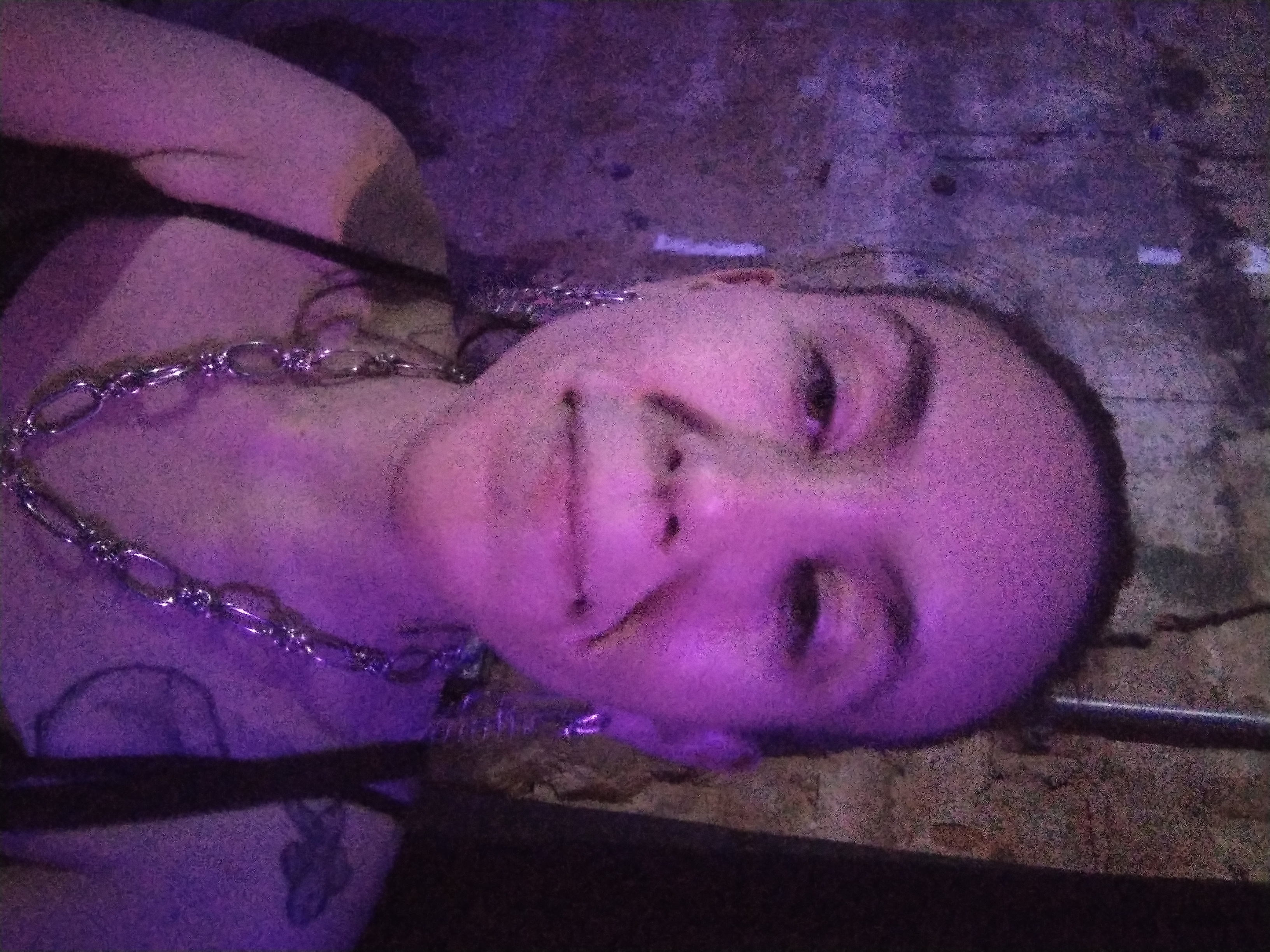 Photo of a white person with their hair pulled back, wearing a chain necklace and earrings, standing in front of a stone wall, in purple lighting.