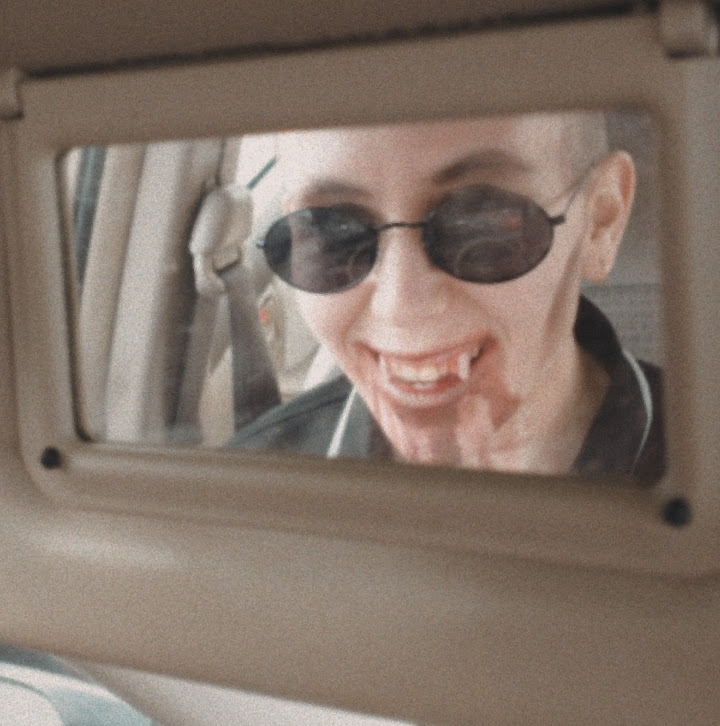 A picture of me smiling and wearing sunglasses and vampire fangs, with blood on their mouth, taken in a car mirror.
